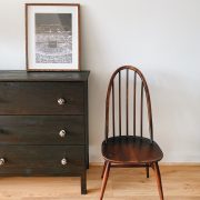 cadeira de madeira e um móvel de madeira com um quadro em cima na frente de uma parede branca em um cômodo com chão de taco